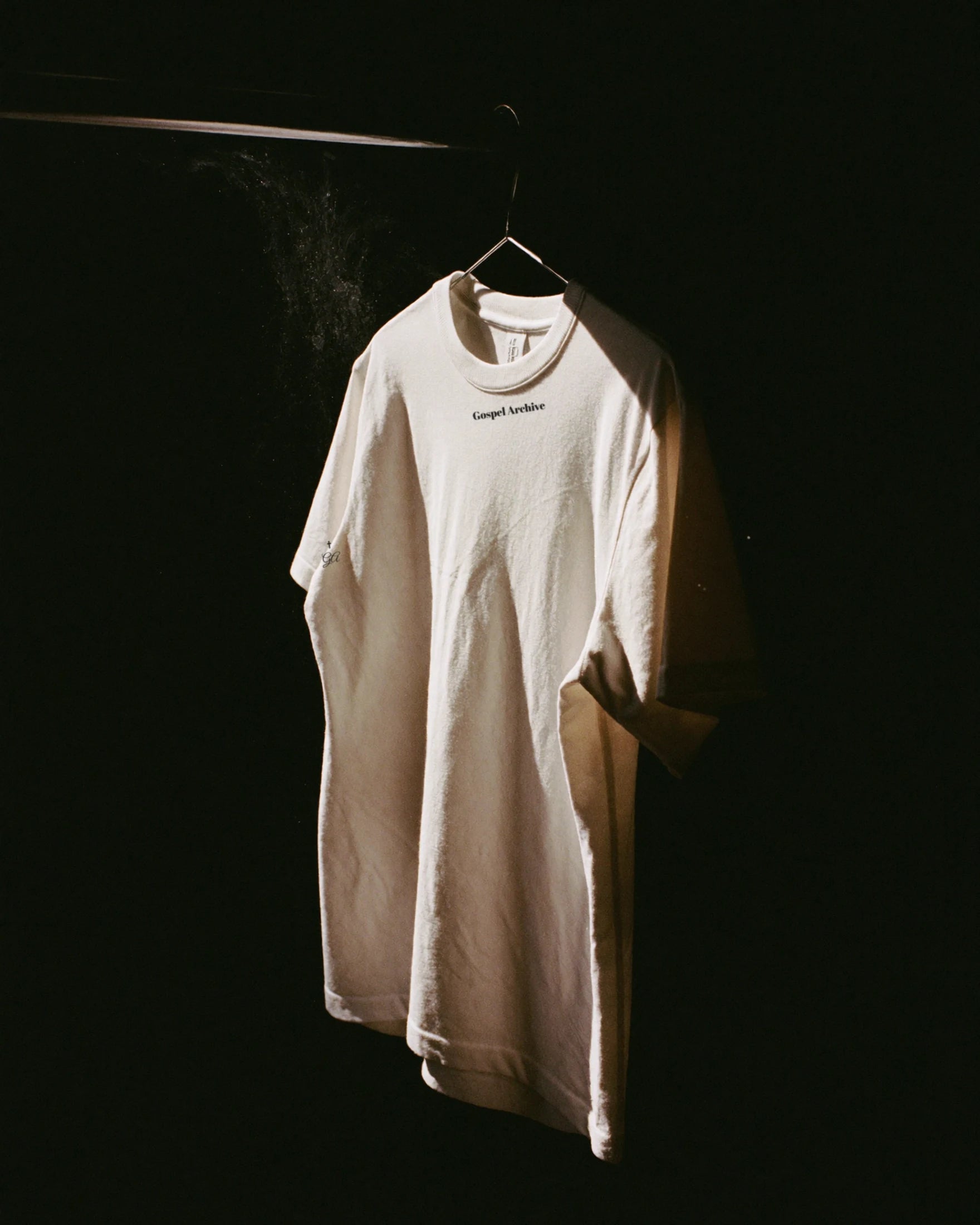 Gospel Archive Drop 001 t-shirt hanging in darkness, lit by a single shaft of light